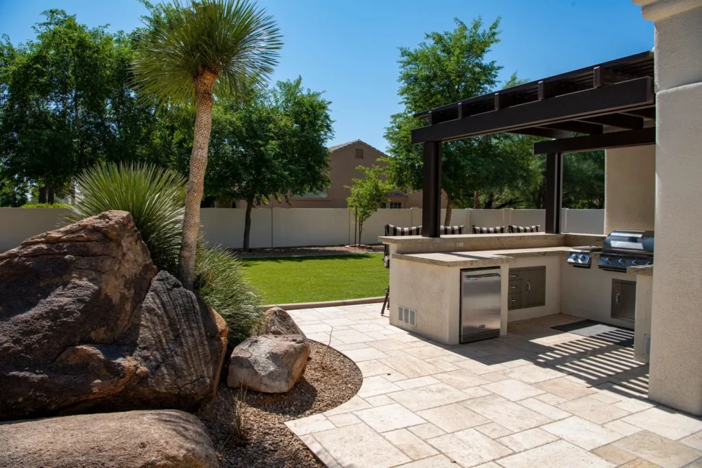 Outdoor kitchen on a paver patio with a wood pergola, palm trees, and landscaped beds
