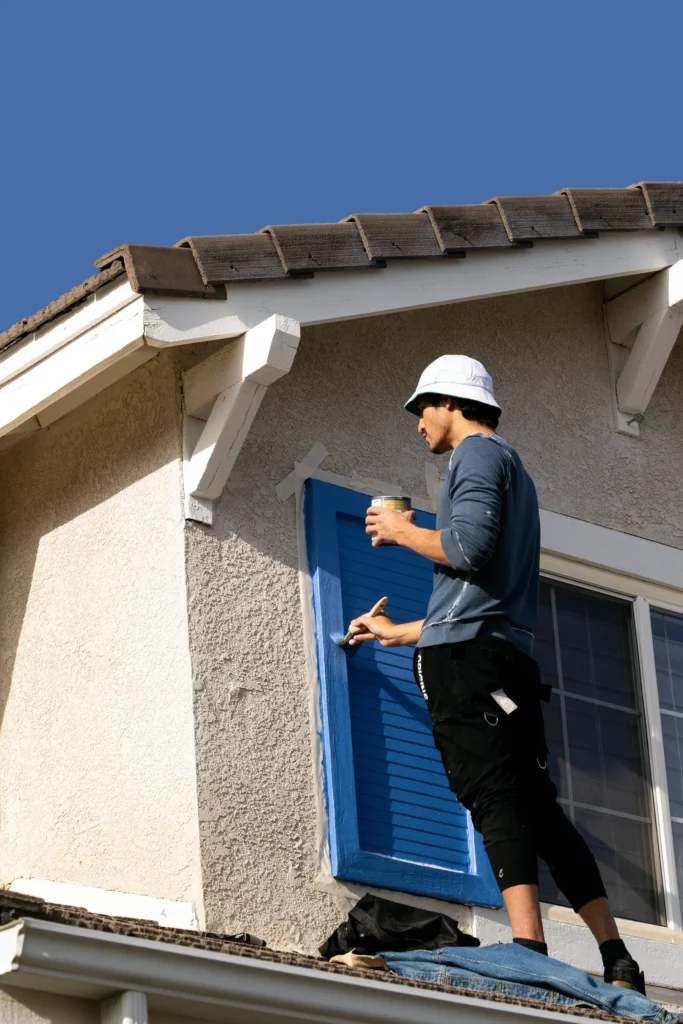 Worker painting a window shutter light blue on the outside of a yellow house